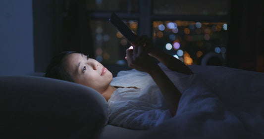 A woman lying in bed using her phone with a soft blue glow from the screen on her face.